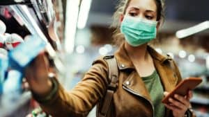 Young woman with face mask using mobile phone and buying groceries in the supermarket during virus pandemic.