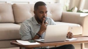 Concentrated young black guy sitting on heated floor at modern coffee table in living room, looking at laptop screen