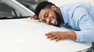 Happy African American Man Hugging New Car In Auto Dealership