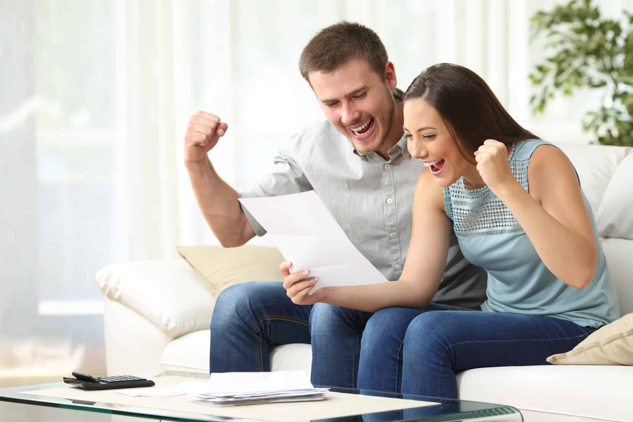 A young couple sitting together on the sofa, reading their financial statements with exuberance.