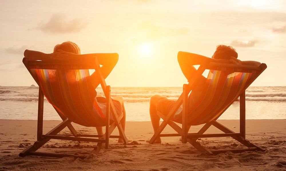 A couple sat on a pair of deck chairs on a beach looking out to sea; the sun is setting on the horizon.