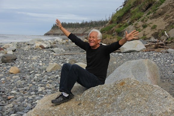 An exuberant, smiling retired man sitting on a rock on a pebble beach, with his arms flung wide open.