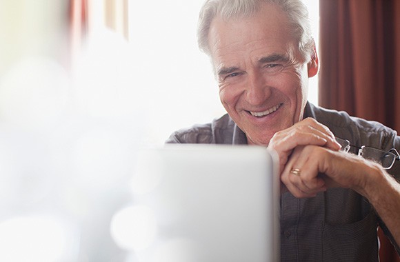 Portrait of smiling senior man holding eyeglasses and using laptop.