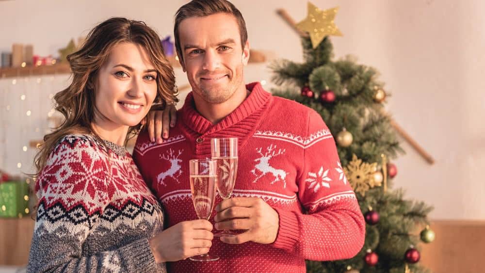smiling couple holding champagne glasses and looking at camera at home with christmas tree