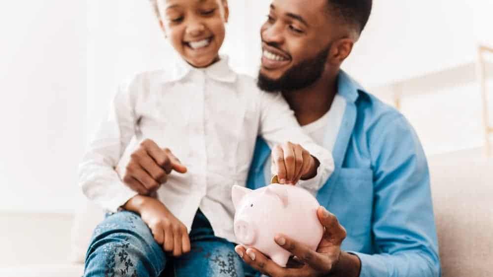 Girl and father putting coin into piggy bank, sitting on sofa at home
