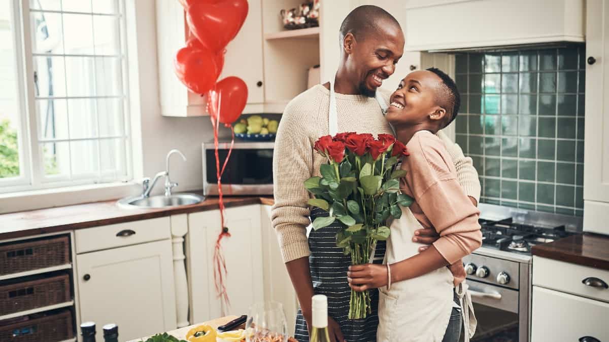 Cropped shot of an affectionate young couple posing with a bunch of flowers in their kitchen on their anniversary