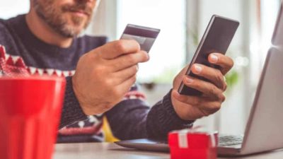 Man sat at laptop computer using credit card to pay online using mobile phone