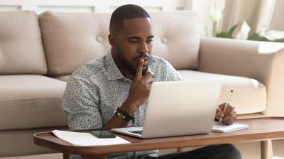 Concentrated young african american black guy sitting on heated floor at modern coffee table in living room, looking at laptop screen
