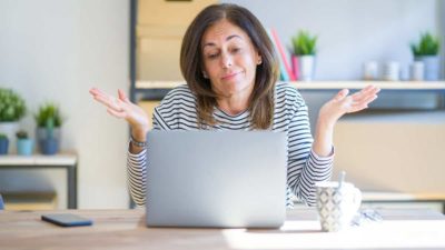 Middle age senior woman sitting at the table at home working using computer laptop clueless and confused expression with arms and hands raised.