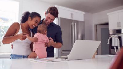 Family With Baby Daughter In Kitchen Using Laptop On Counter