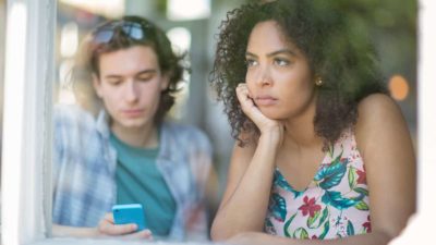 Frustrated young woman gazing out of the window while her male companion stares at his phone