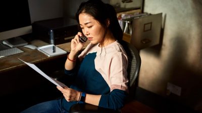 Serious mid adult Asian woman listening on phone, sitting in office chair in dark room, holding paperwork