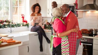 Multi-ethnic adult family celebrating with champagne, talking and dancing in the kitchen while preparing dinner on Christmas Day