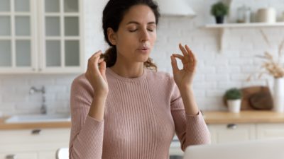 Mindful young woman breathing out with closed eyes, calming down in stressful situation, working on computer in modern kitchen.