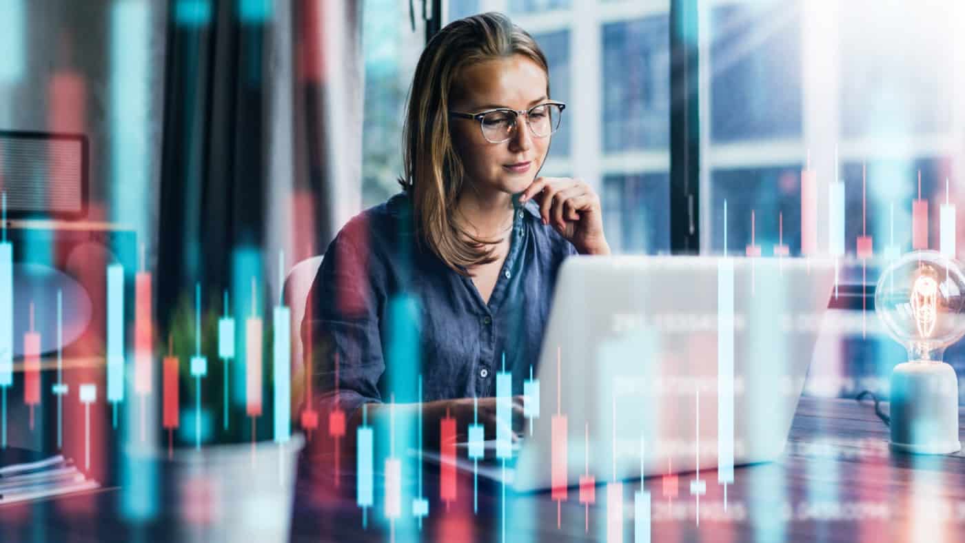 現代のオフィスで働く若い女性。 技術的な価格グラフとインジケーター、赤と緑のローソク足チャート、株式取引のコンピューター画面の背景。