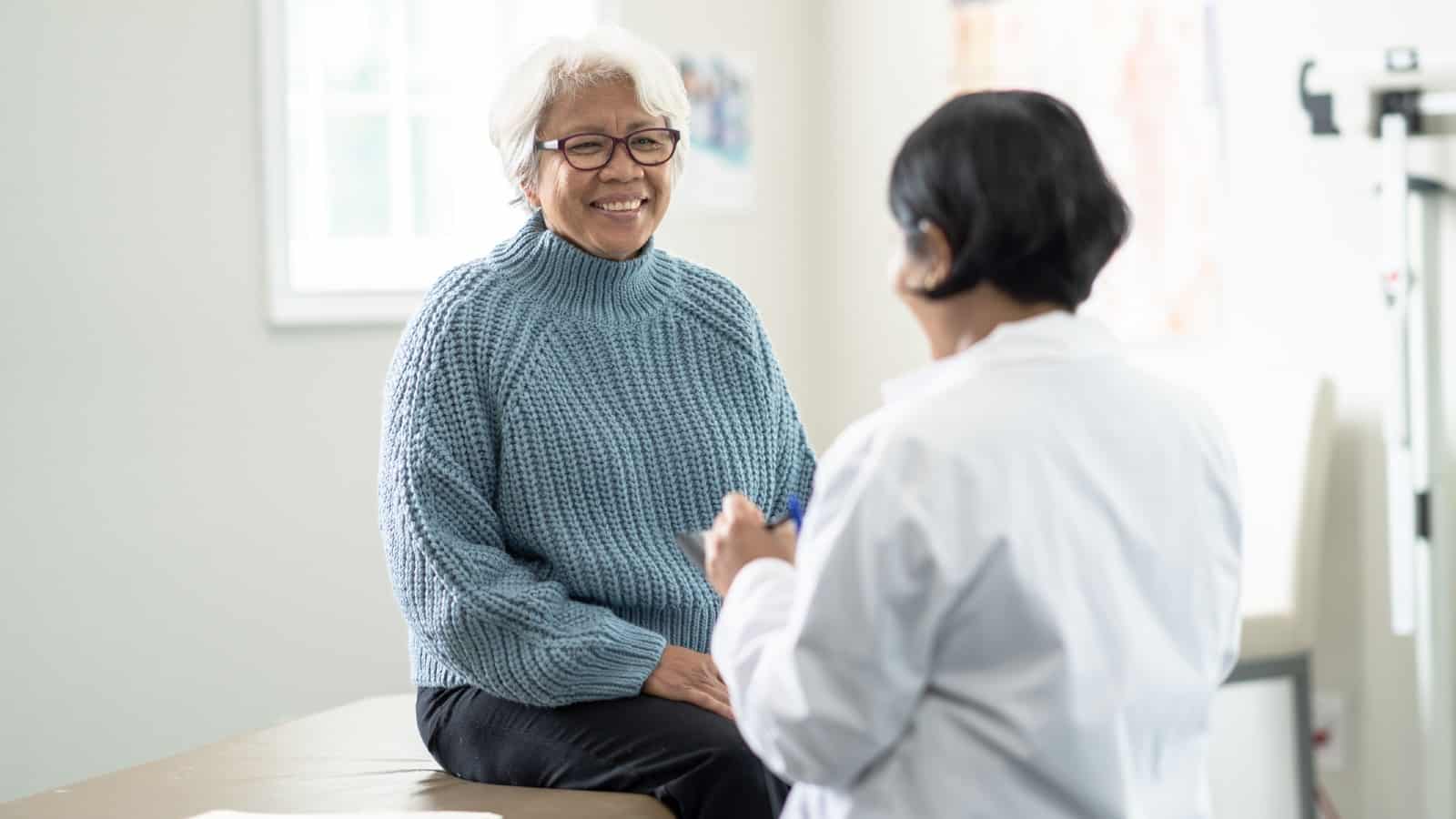 A senior woman sits up on the exam table at a doctors appointment. She is dressed casually in a blue sweater and has a smile on her face as she glances at the doctor. Her female doctor is wearing a white lab coat and seated in front of her as she takes notes on a tablet.
