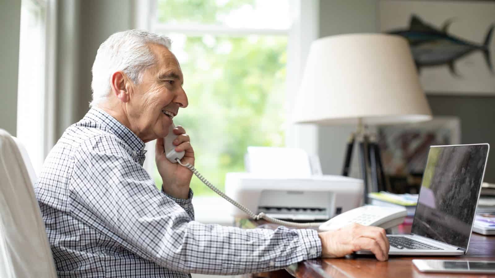 Smiling senior white man talking through telephone while using laptop at desk.