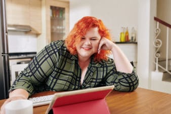 Happy young plus size woman sitting at kitchen table and watching tv series on tablet computer