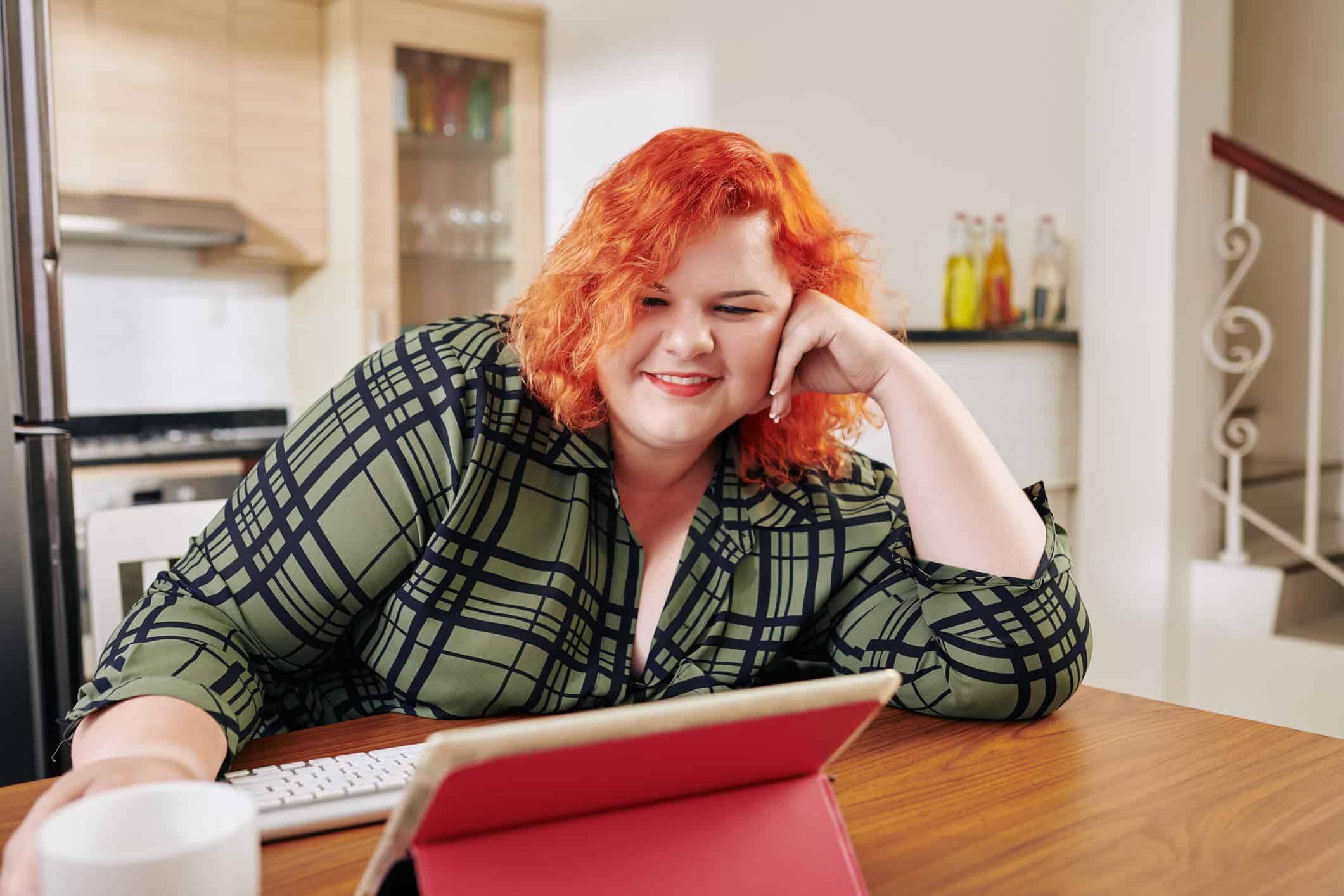 Ευτυχισμένη νεαρή γυναίκα σε μεγάλο μέγεθος που κάθεται στο τραπέζι της κουζίνας και παρακολουθεί τηλεοπτικές σειρές σε ένα tablet