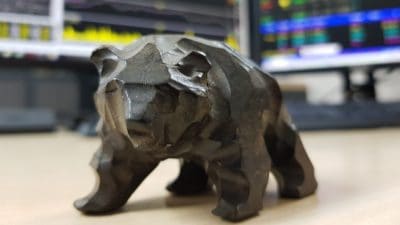 Tabletop model of a bear sat on desk in front of monitors showing stock charts