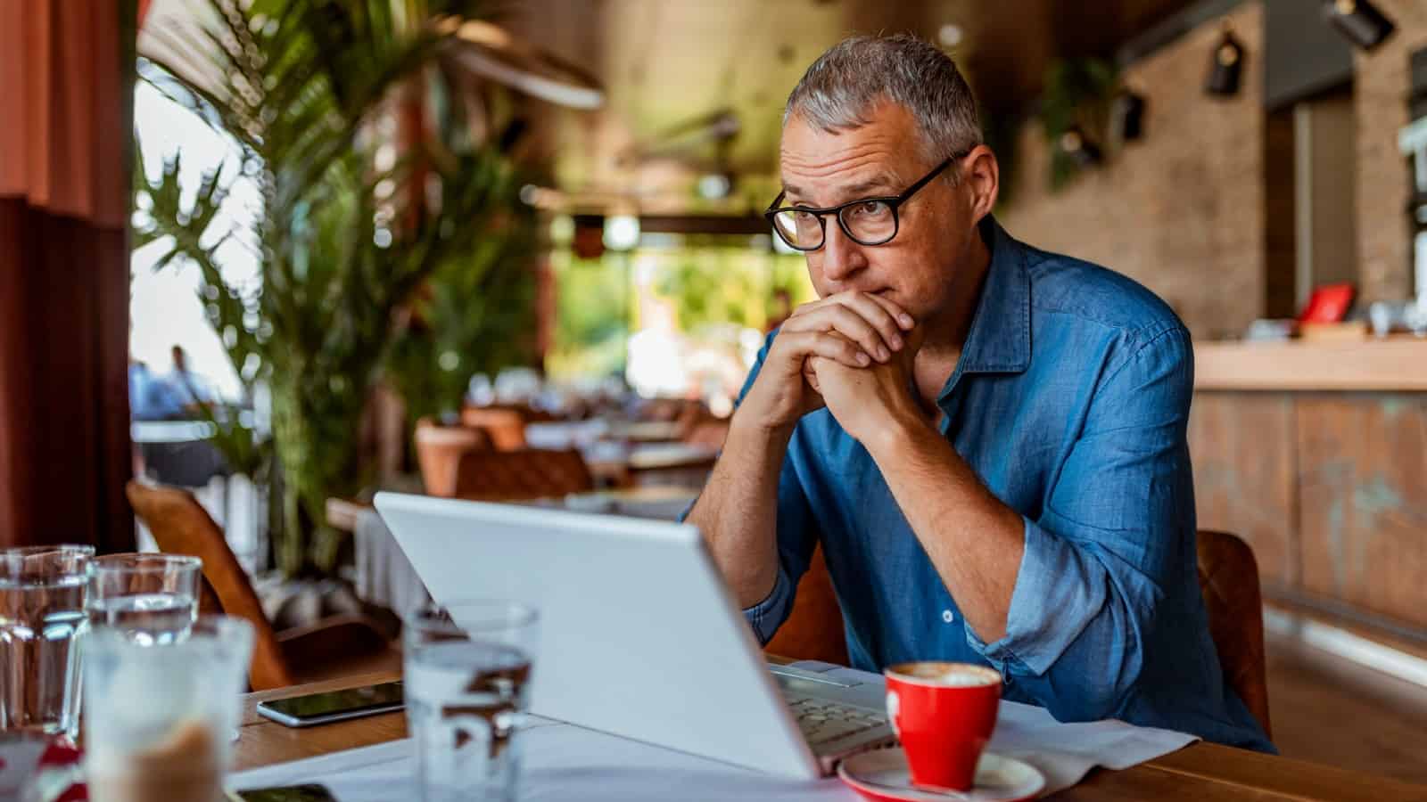 Middle-aged white man wearing glasses, staring into space over the top of his laptop in a coffee shop