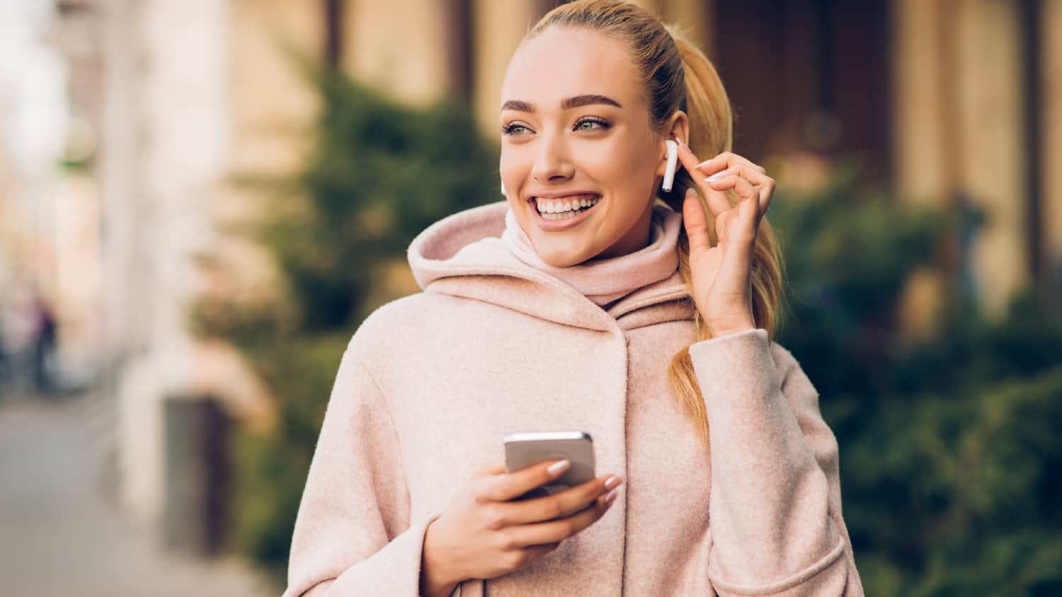 कान में एयरपॉड्स लगाए आईफोन पकड़े हुए मुस्कुराती हुई सफेद महिला