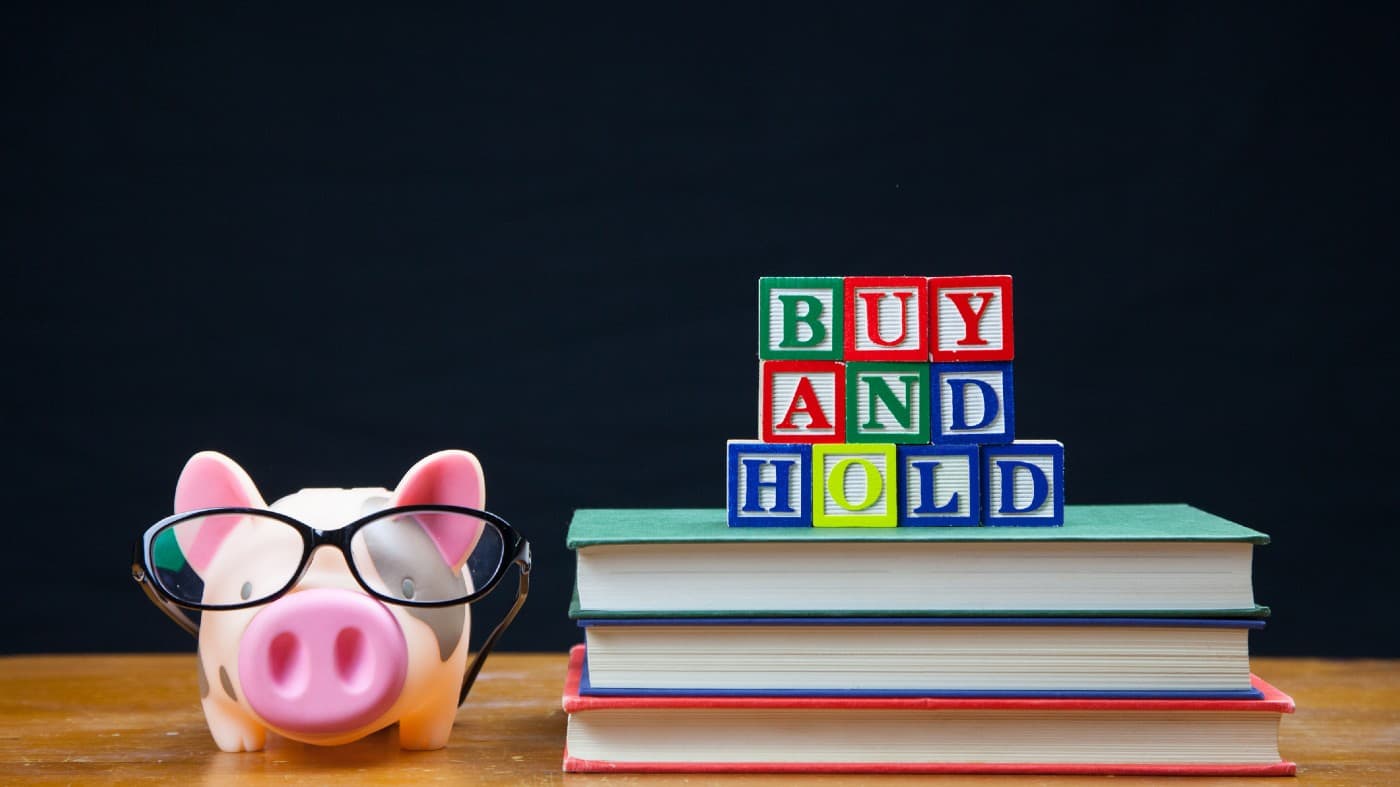 BUY AND HOLD spelled in letters on top of a pile of books. Alongside is a piggy bank in glasses. Buy and hold is a popular long term stock and shares strategy.