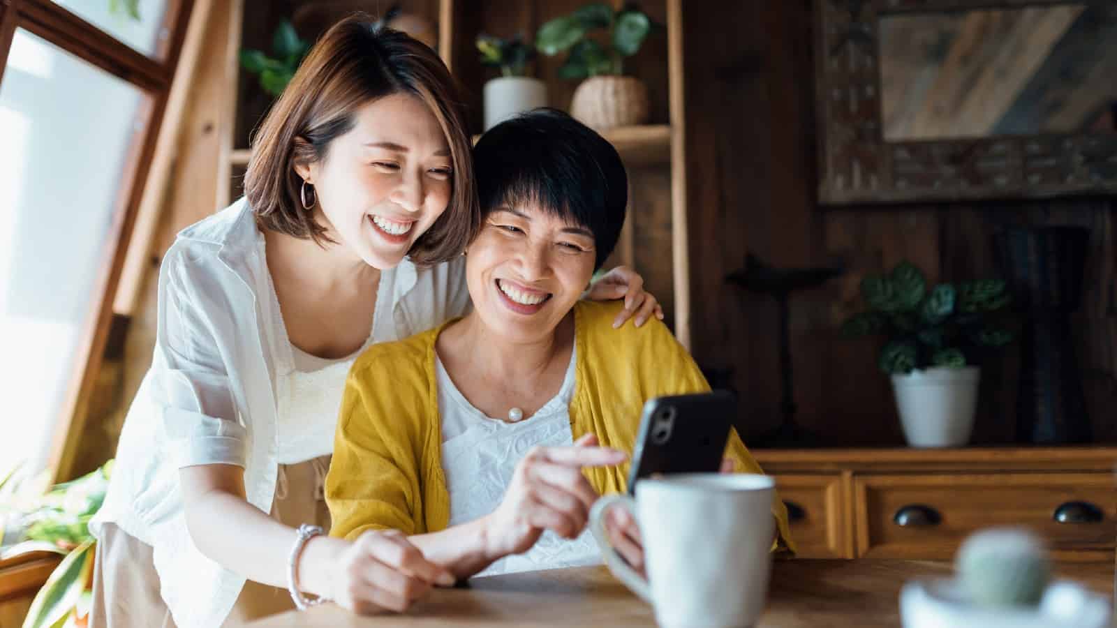 Affectionate Asian senior mother and daughter using smartphone together at home, smiling joyfully