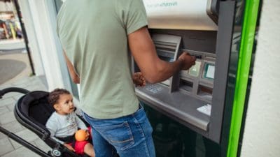 Man putting his card into an ATM machine while his son sits in a stroller beside him.