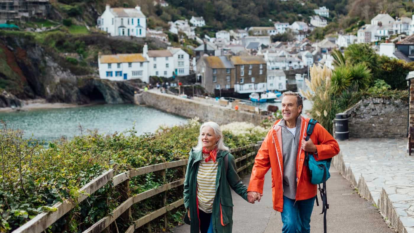 A senior man and his wife holding hands walking up a hill on a footpath looking away from the camera at the view. The fishing village of Polperro is behind them.