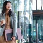 Young Asian woman holding a cup of takeaway coffee and folders containing paperwork, on her way into the office