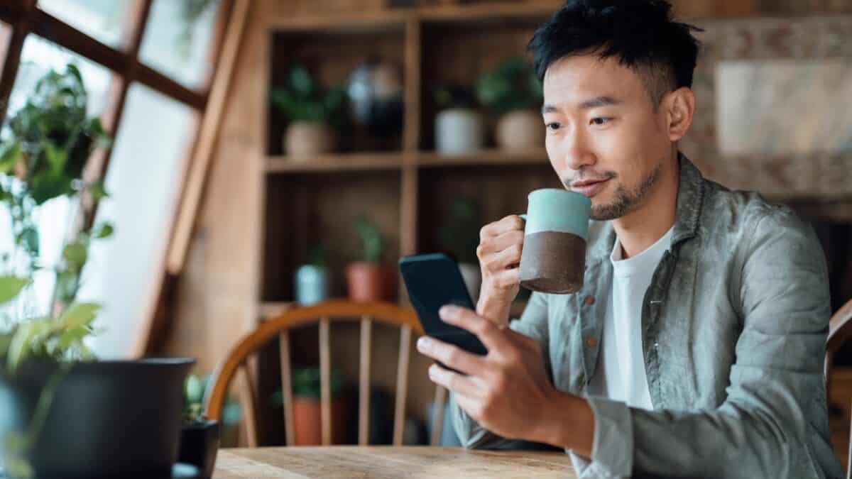 Νεαρός Ασιάτης πίνει καφέ στο σπίτι και κοιτάζει το τηλέφωνό του