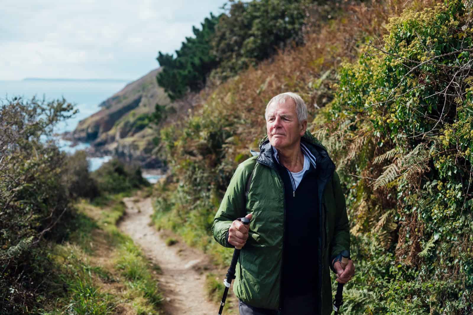 A senior man using hiking poles, on a hike on a coastal path along the coastline of Cornwall.