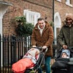 Two male friends are out in Tynemouth, North East UK. They are walking on a sidewalk and pushing their baby sons in strollers. They are wearing warm clothing.