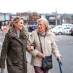 A mature woman help a senior woman out of a car as she takes her to the shops.